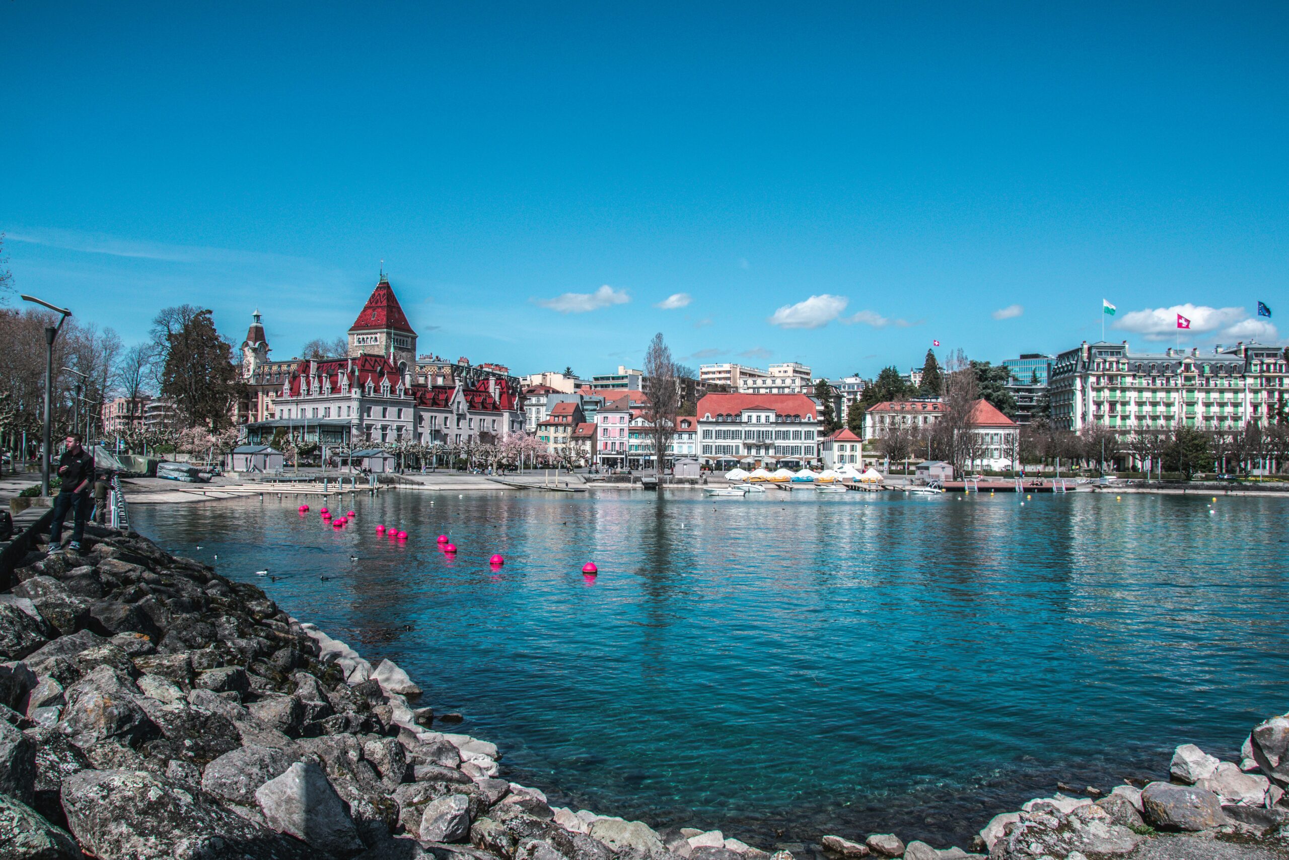 A scenic view of a waterfront in Lausanne, Switzerland. The image shows a clear blue sky with a few clouds, and the calm waters of Lake Geneva reflecting the buildings along the shore. Prominent structures include the Château d'Ouchy with its red roof and tower, surrounded by other historic buildings and modern establishments. Pink buoys float in the water near the shore, and there are boats docked at a marina to the right. In the foreground, there is a rocky shoreline with two people standing on it.