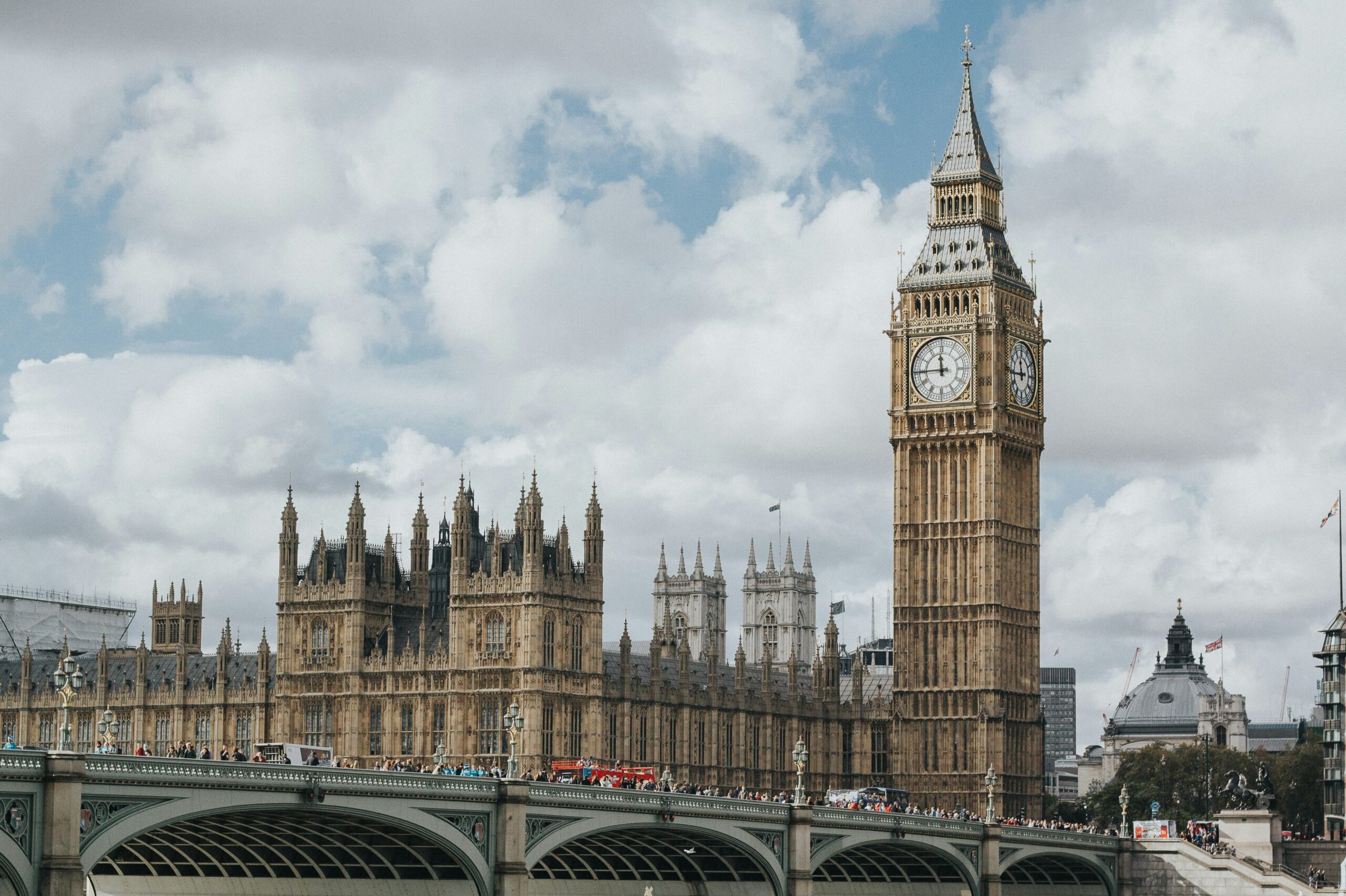 The image shows the iconic Big Ben clock tower and the Houses of Parliament in London, England. The sky is filled with clouds, and Westminster Bridge can be seen in the foreground with people and vehicles crossing it.