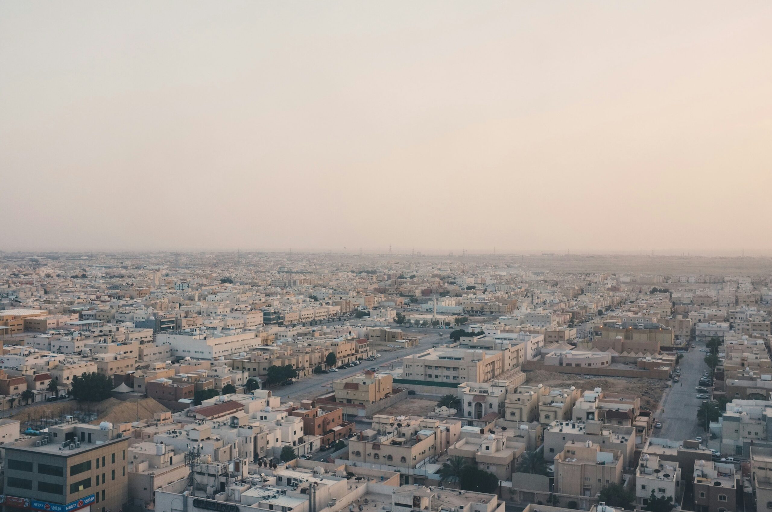 Aerial view of a sprawling cityscape with numerous beige and white buildings under a hazy sky. The buildings are closely packed together, with some trees and roads visible between them. The horizon is barely visible due to the haze.