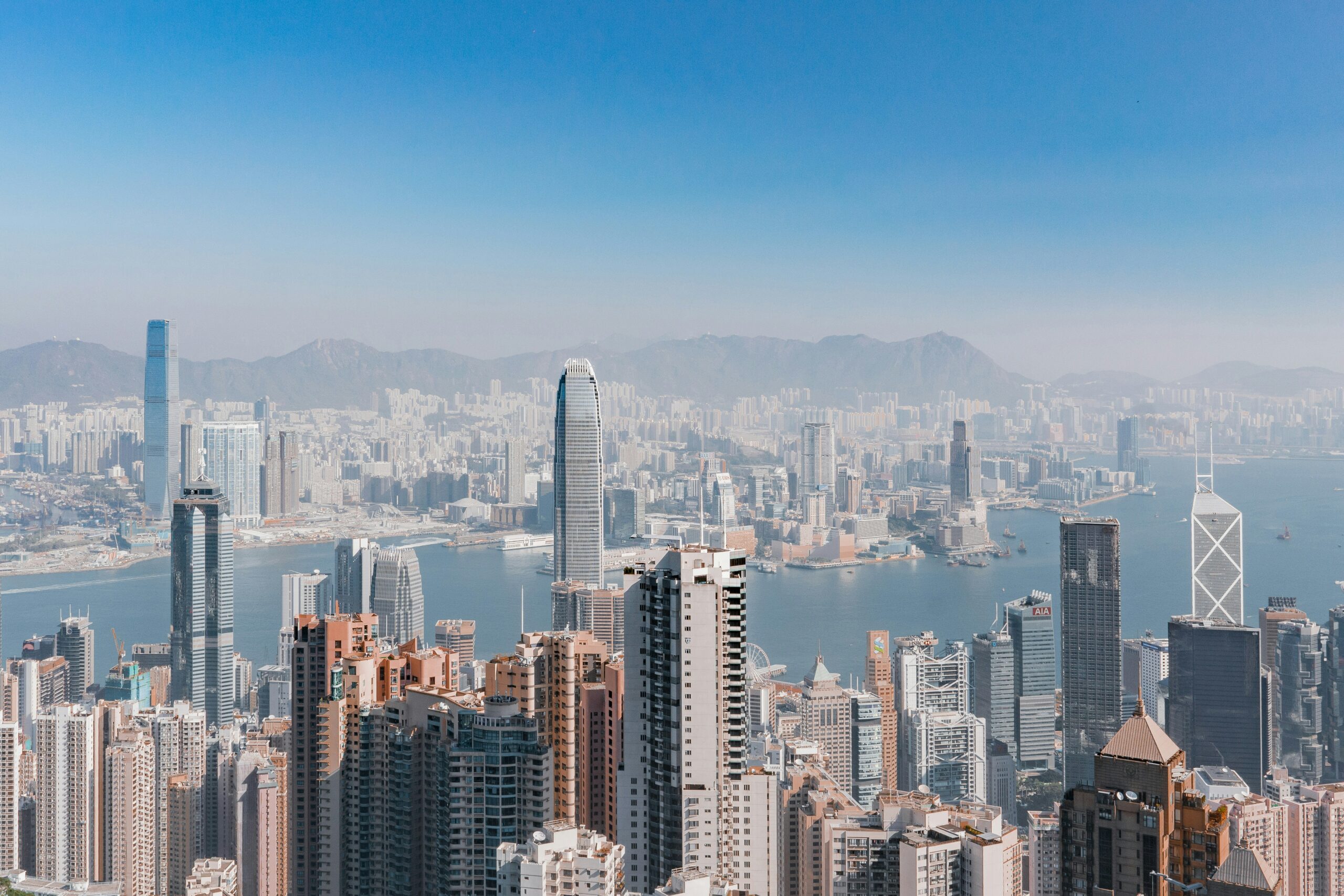 A panoramic view of Hong Kong's skyline featuring numerous high-rise buildings, with Victoria Harbour and distant mountains in the background under a clear blue sky.