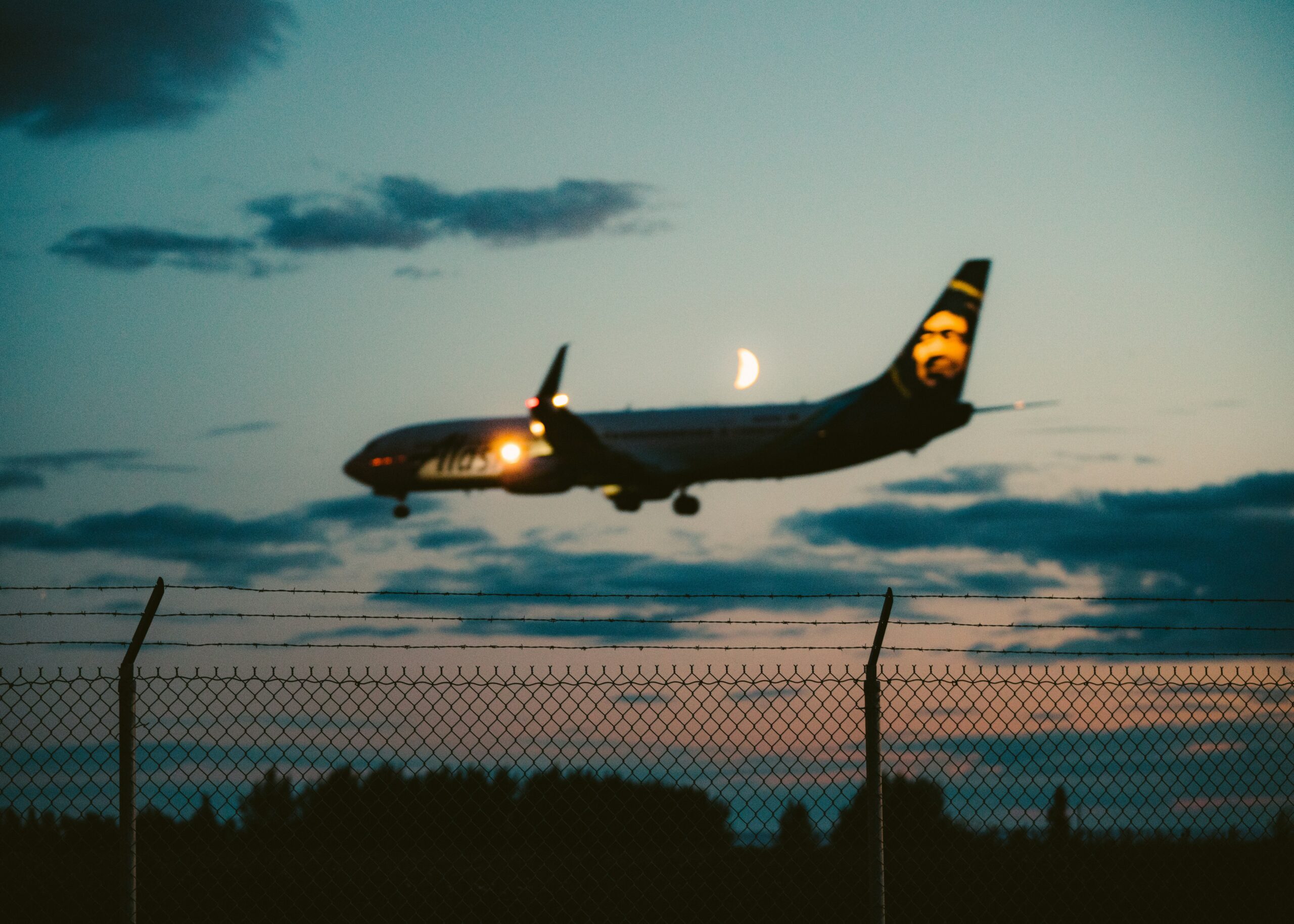 An airplane is captured in mid-flight during dusk, with its landing lights on and a crescent moon visible in the background. The aircraft appears to be descending towards an airport, as indicated by the barbed wire fence in the foreground. The sky is a gradient of blue and orange hues, with scattered clouds adding to the serene atmosphere.