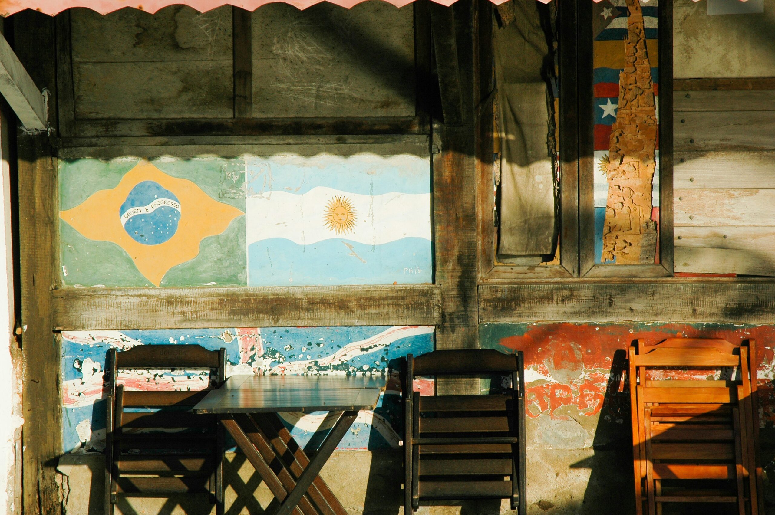 A weathered wooden wall features painted flags of Brazil, Argentina, and Venezuela. Below the flags, there is a small wooden table with two dark wooden chairs on either side and one light wooden chair to the right.