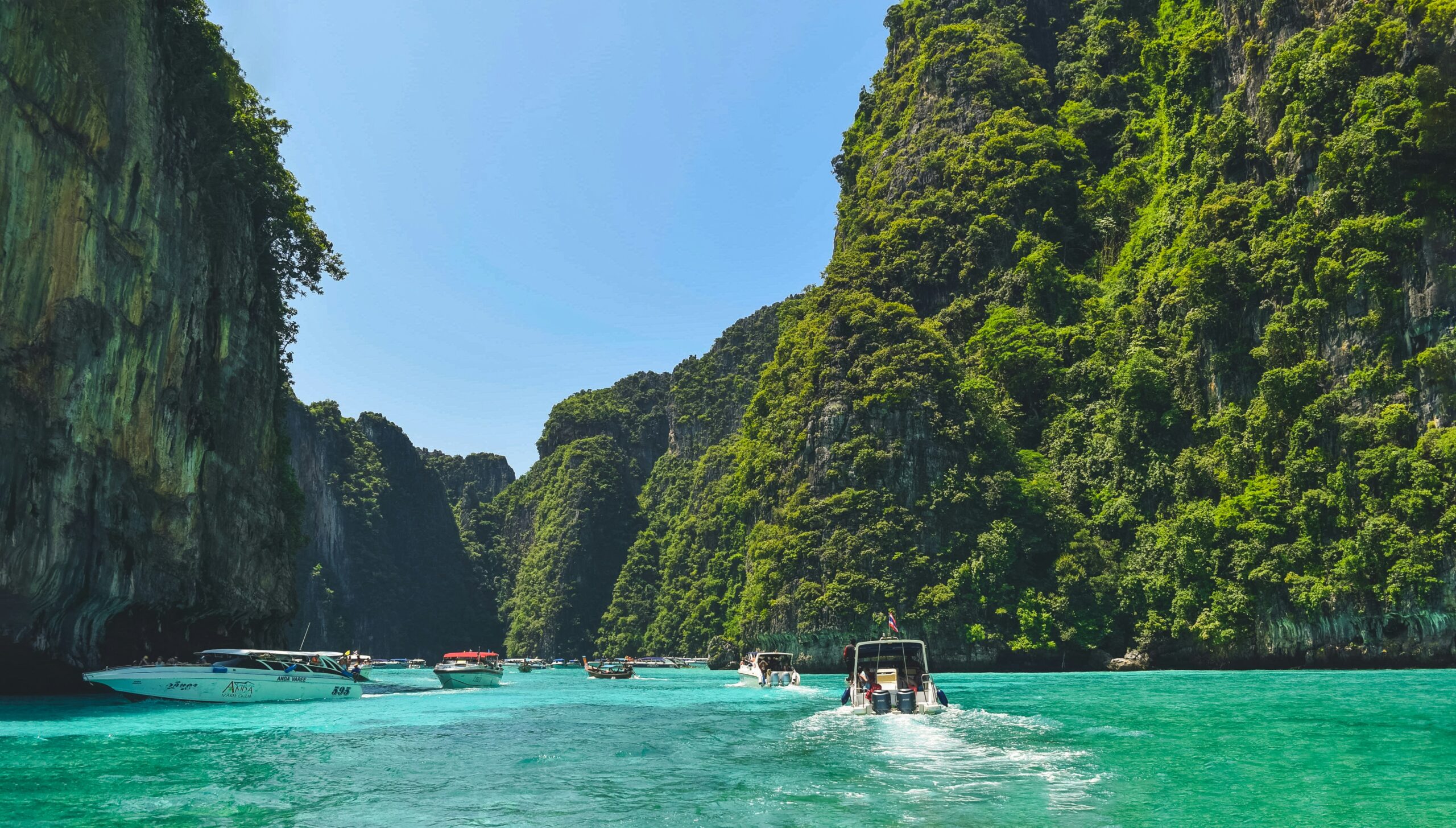 A scenic view of a tropical bay surrounded by towering limestone cliffs covered in lush greenery. Several boats are seen cruising on the clear turquoise waters, with one boat in the foreground creating a trail of white foam behind it.
