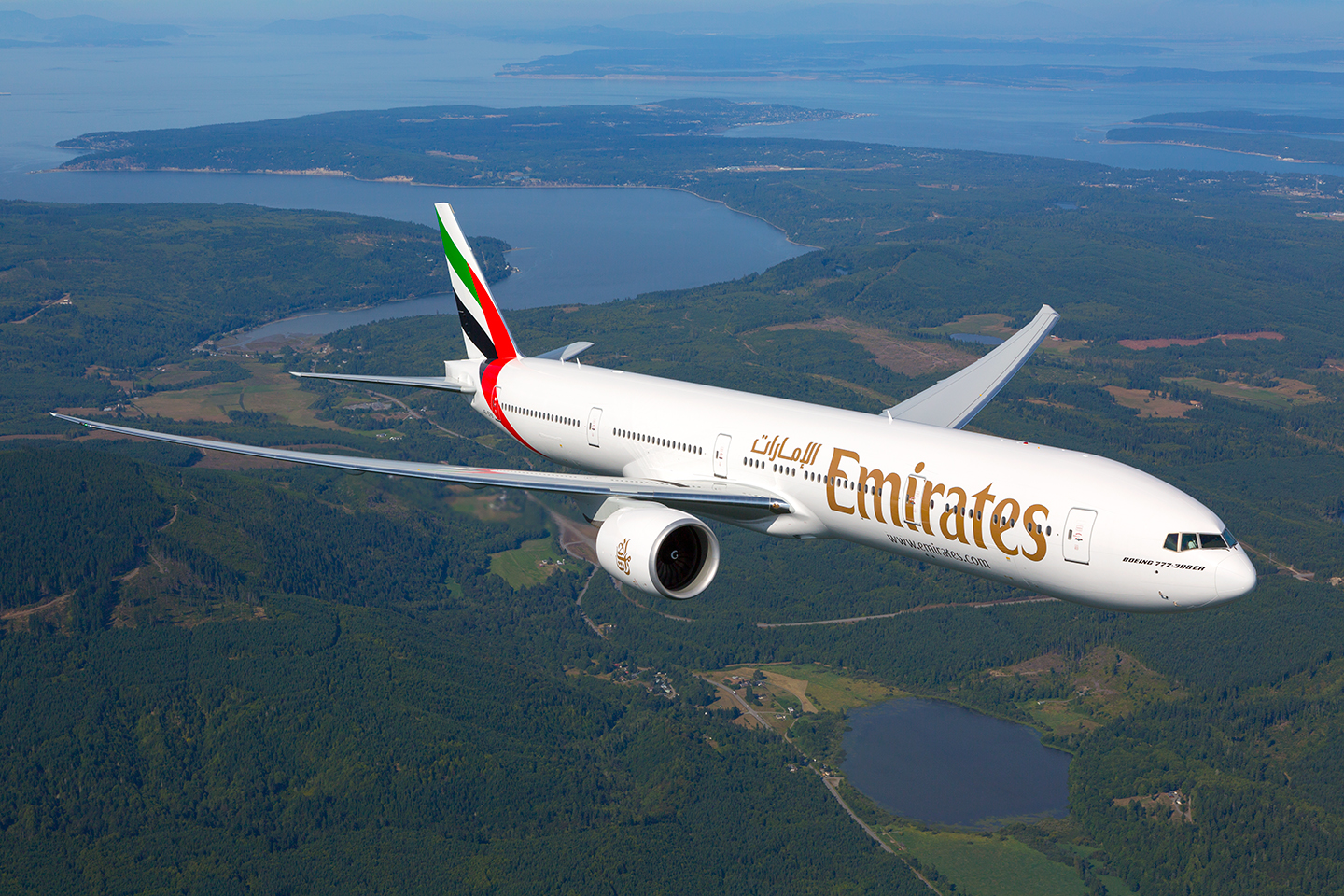 An Emirates Boeing 777-300ER aircraft is flying over a lush, green landscape with several bodies of water visible below. The airplane's white fuselage features the Emirates logo in gold lettering, and the tail fin displays the airline's red, green, black, and white colors.