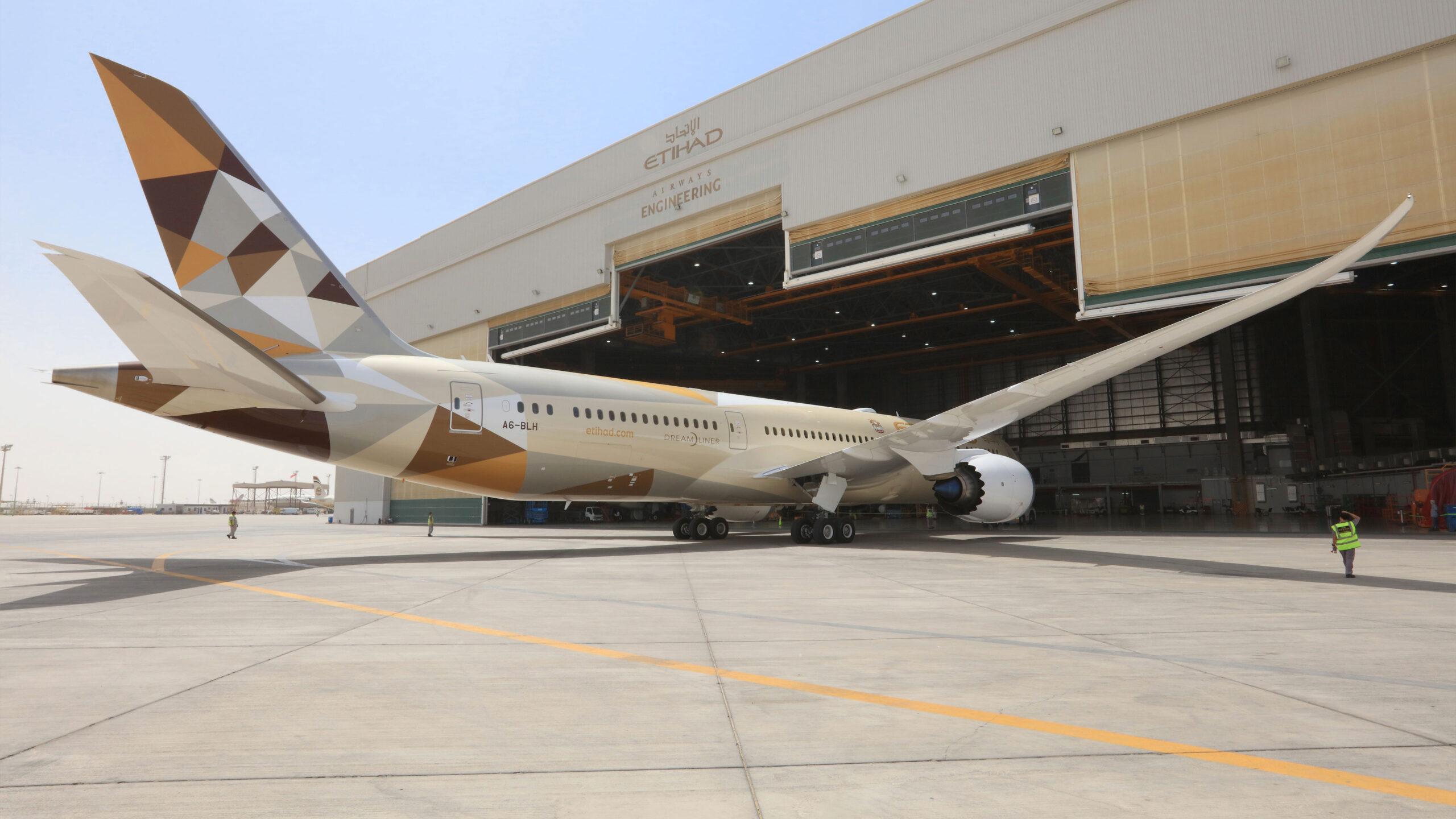 A large Etihad Airways aircraft, specifically a Boeing 787 Dreamliner with the registration number A6-BLH, is parked outside an Etihad Airways Engineering hangar. The airplane features a distinctive geometric livery in shades of brown, gold, and white. The hangar doors are open, revealing part of the interior. Two ground crew members in high-visibility vests are visible near the aircraft.