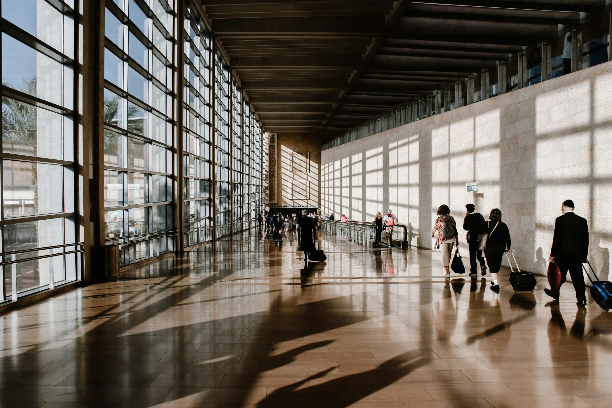 A group of people with luggage walk through a bright, spacious airport corridor with large windows and sunlight streaming in, creating long shadows on the shiny floor.