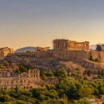 A panoramic view of the Acropolis in Athens, Greece, during sunset. The Parthenon stands prominently on the right side of the image, partially covered with scaffolding for restoration. Other ancient structures such as the Erechtheion and the Odeon of Herodes Atticus are also visible. The surrounding area is lush with greenery and modern buildings can be seen at the base.