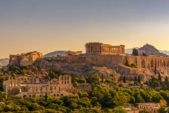 A panoramic view of the Acropolis in Athens, Greece, during sunset. The Parthenon stands prominently on the right side of the image, partially covered with scaffolding for restoration. Other ancient structures such as the Erechtheion and the Odeon of Herodes Atticus are also visible. The surrounding area is lush with greenery and modern buildings can be seen at the base.