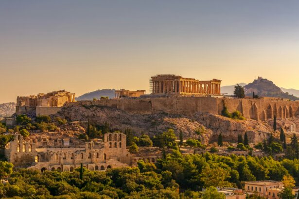 A panoramic view of the Acropolis in Athens, Greece, during sunset. The Parthenon stands prominently on the right side of the image, partially covered with scaffolding for restoration. Other ancient structures such as the Erechtheion and the Odeon of Herodes Atticus are also visible. The surrounding area is lush with greenery and modern buildings can be seen at the base.
