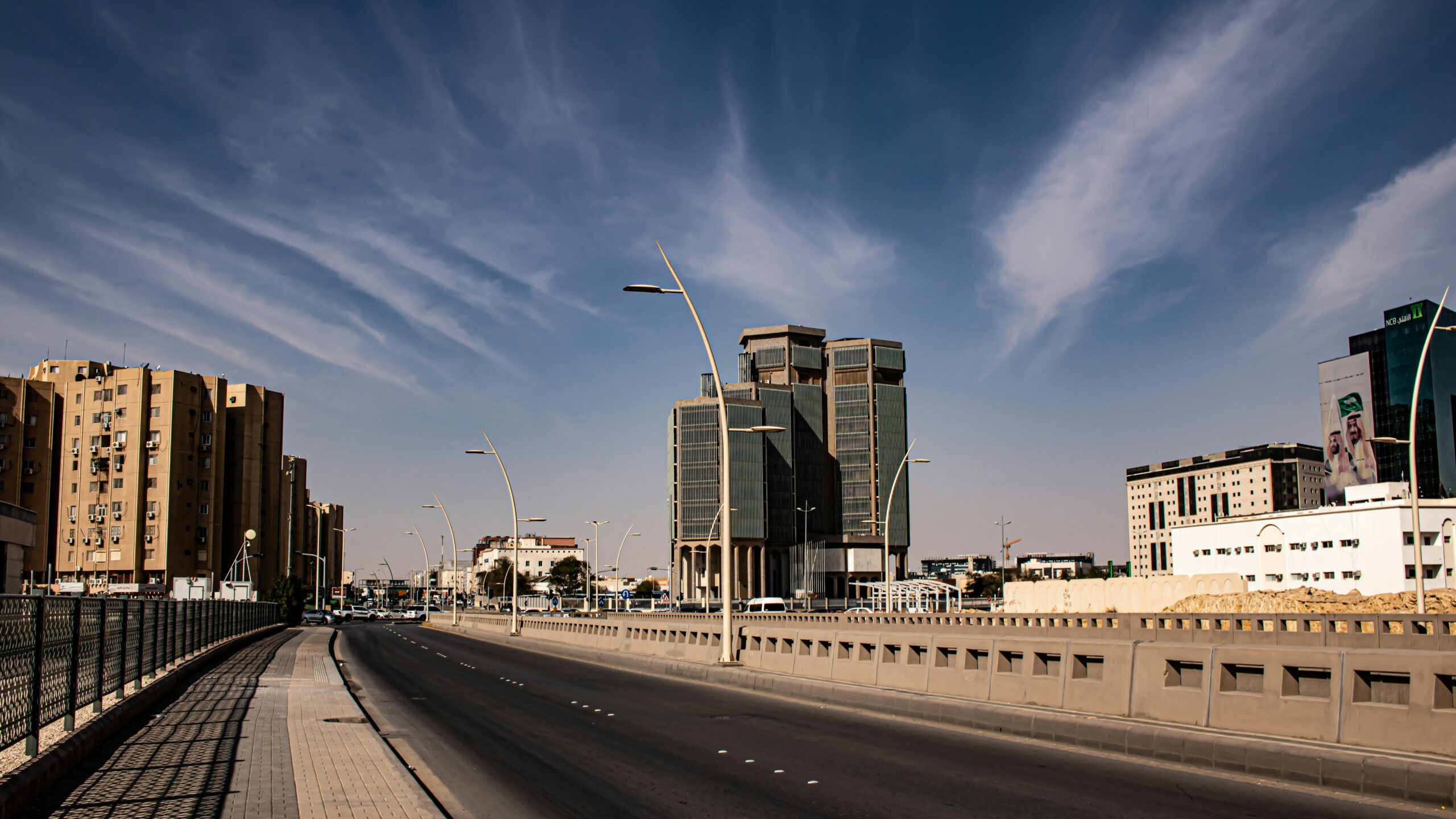 A wide-angle view of a city street with modern buildings on both sides. The street is lined with tall, uniquely designed street lamps and has minimal traffic. On the left side, there are beige residential buildings, while on the right side, there are taller commercial buildings with glass facades. The sky above is clear with some wispy clouds.