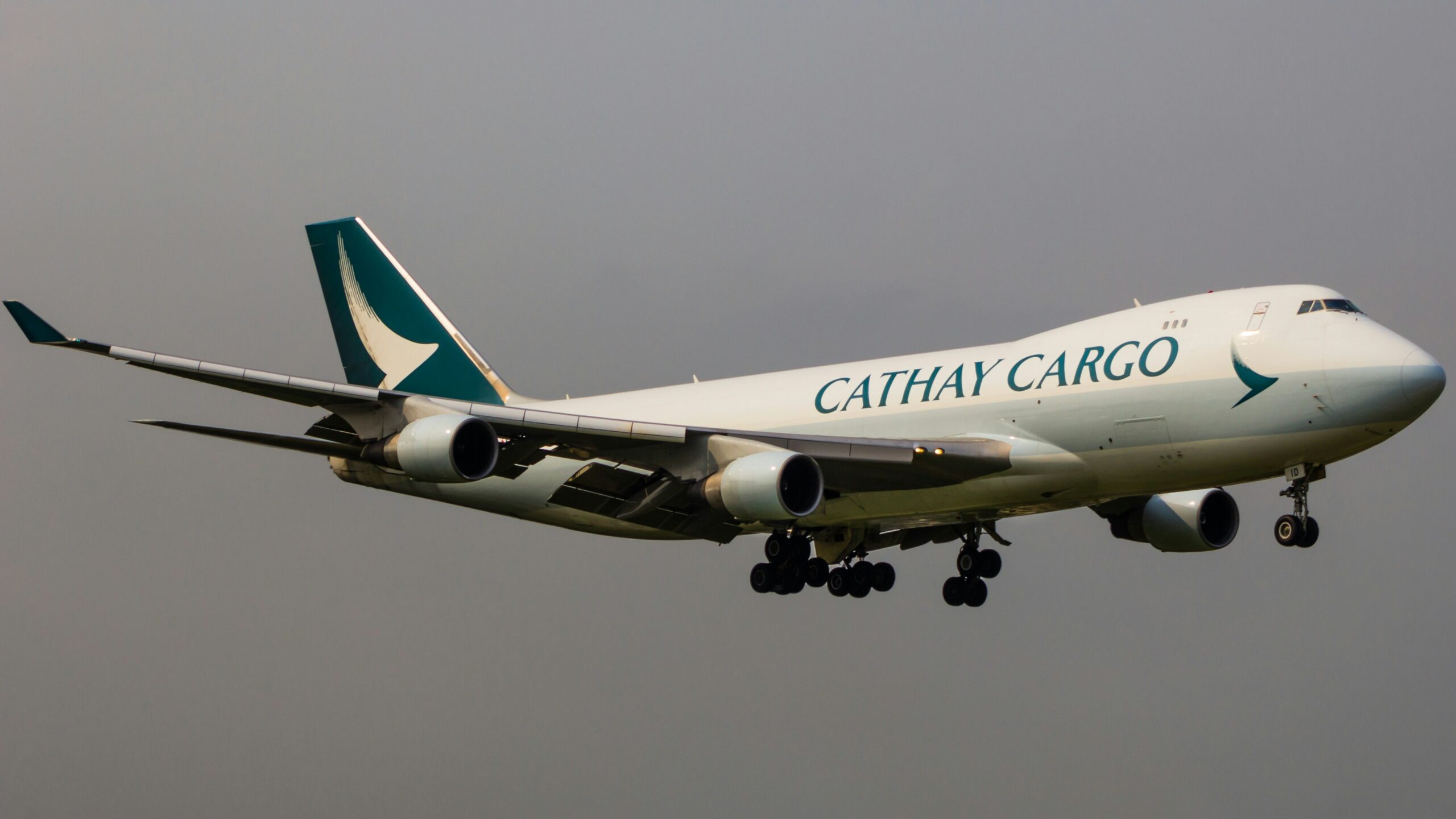 A Cathay Cargo aircraft is captured in mid-air during its landing approach. The aeroplane features the airline's distinctive green and white livery with the Cathay Cargo logo prominently displayed on the fuselage.