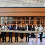 A group of eleven people, including airline staff and officials, stand in a line holding a ribbon during a ceremonial event at an airport lounge. The ribbon features the Cathay Pacific logo and Chinese characters. The background shows a modern lounge area with glass windows and wooden panels.