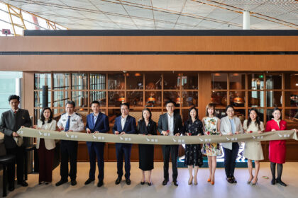 A group of eleven people, including airline staff and officials, stand in a line holding a ribbon during a ceremonial event at an airport lounge. The ribbon features the Cathay Pacific logo and Chinese characters. The background shows a modern lounge area with glass windows and wooden panels.