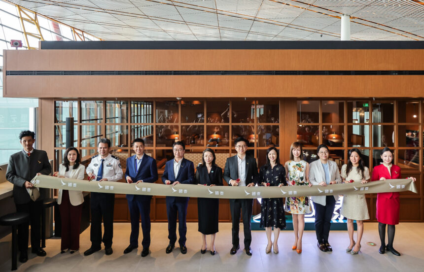 A group of eleven people, including airline staff and officials, stand in a line holding a ribbon during a ceremonial event at an airport lounge. The ribbon features the Cathay Pacific logo and Chinese characters. The background shows a modern lounge area with glass windows and wooden panels.