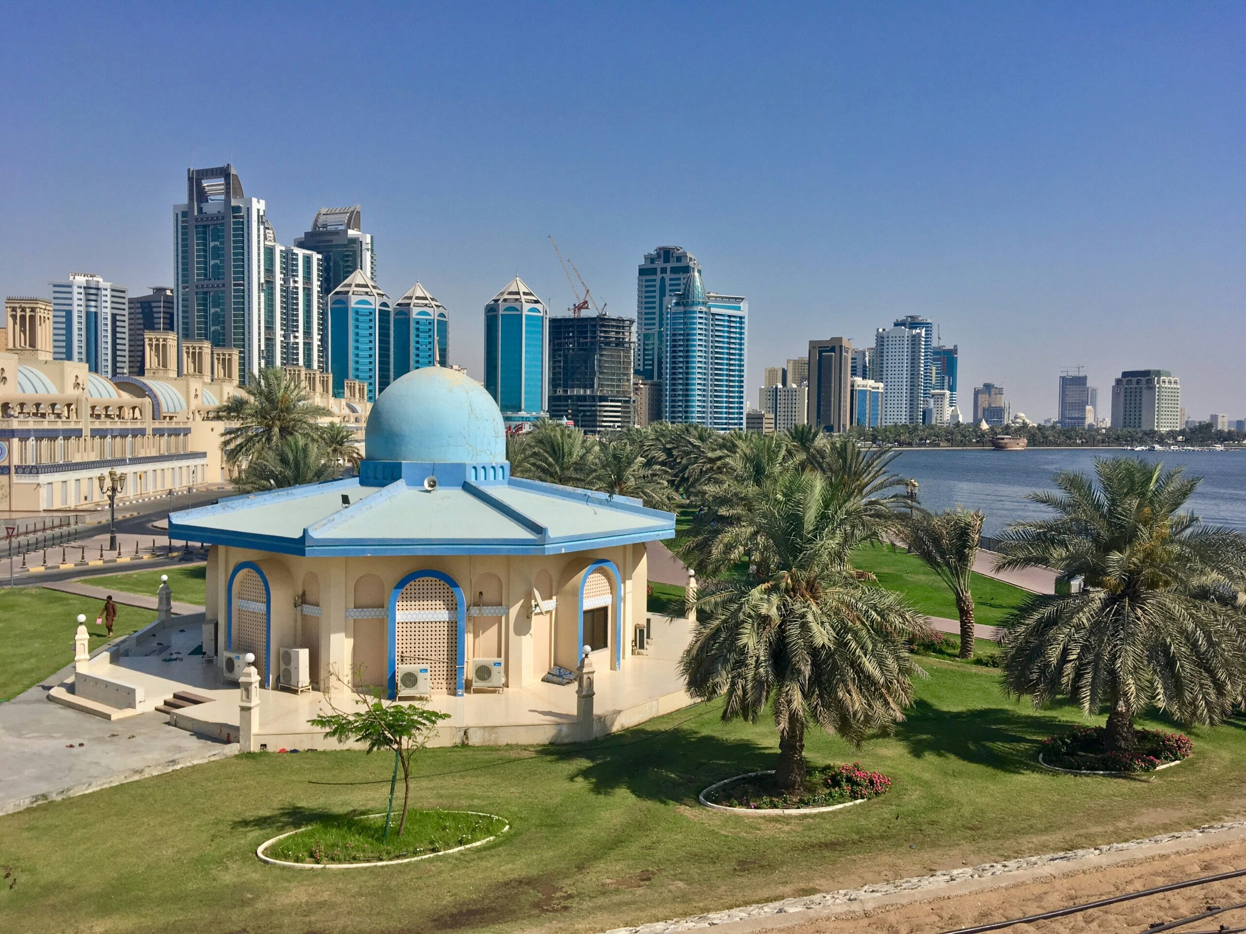 A scenic view of a cityscape featuring a mix of modern high-rise buildings and traditional architecture. In the foreground, there is a small building with a blue dome roof surrounded by palm trees and green lawns. The background showcases tall skyscrapers with glass facades, set against a clear blue sky. A body of water runs alongside the city, adding to the picturesque setting.