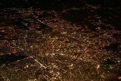An aerial view of a city at night, showcasing a vast network of illuminated streets and buildings. The city lights form intricate patterns against the dark backdrop, highlighting the urban landscape.