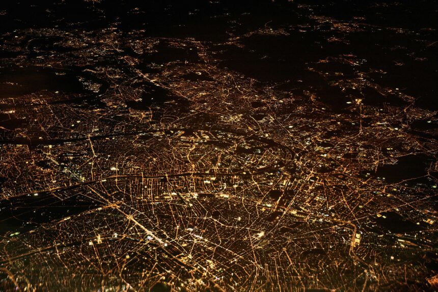 An aerial view of a city at night, showcasing a vast network of illuminated streets and buildings. The city lights form intricate patterns against the dark backdrop, highlighting the urban landscape.