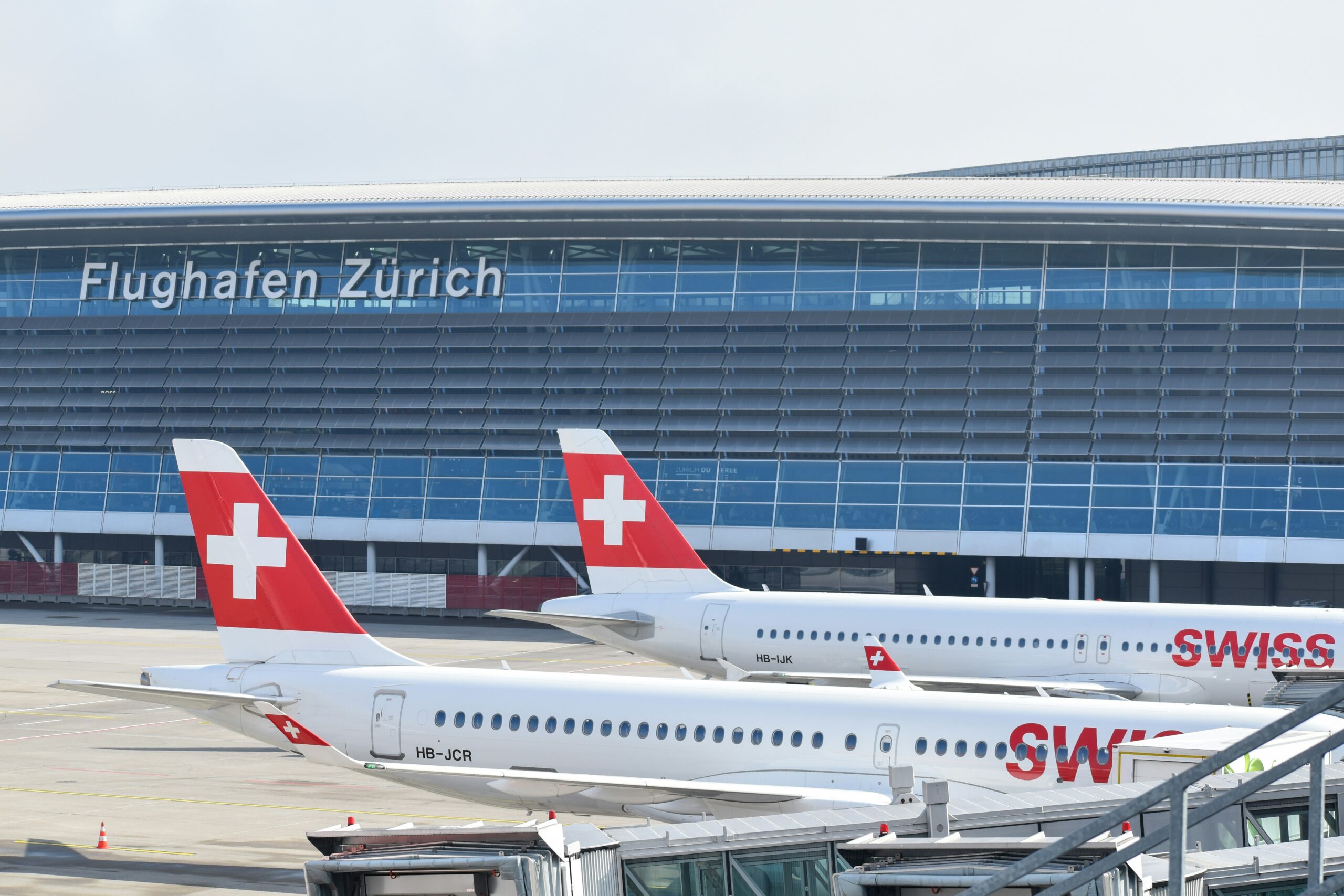 Three Swiss International Air Lines aircraft are parked at Zurich Airport, with the airport terminal building in the background. The terminal has a large sign that reads "Flughafen Zürich" in white letters on a blue facade.