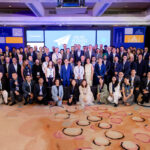 A large group of people, dressed in business attire, are gathered for a group photo at the Airline Business Seminar. The background features a blue and white banner with the text "AIRLINE BUSINESS SEMINAR" and the logo of Embraer. The setting appears to be an indoor conference room with colorful carpeting.
