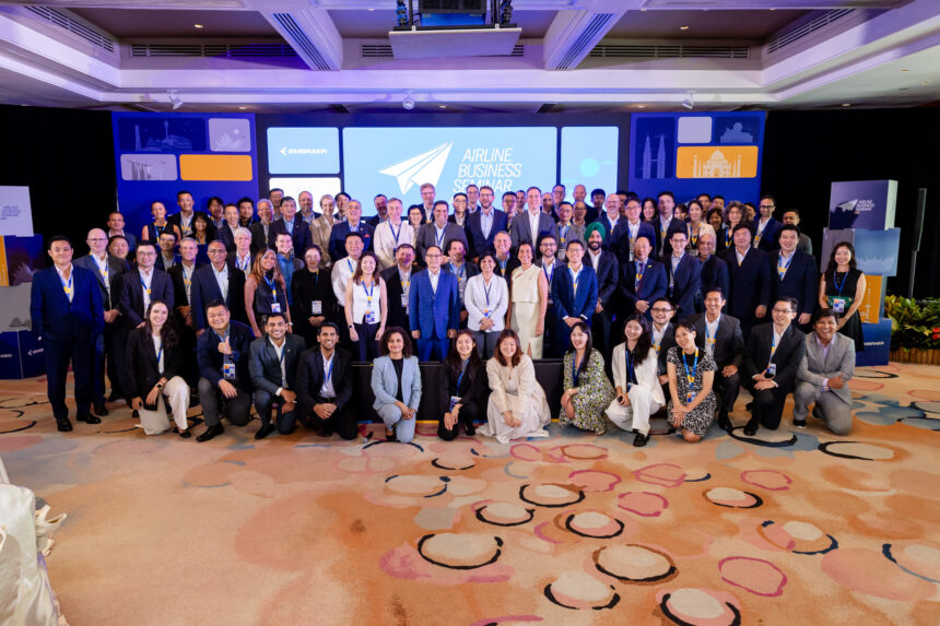 A large group of people, dressed in business attire, are gathered for a group photo at the Airline Business Seminar. The background features a blue and white banner with the text "AIRLINE BUSINESS SEMINAR" and the logo of Embraer. The setting appears to be an indoor conference room with colorful carpeting.