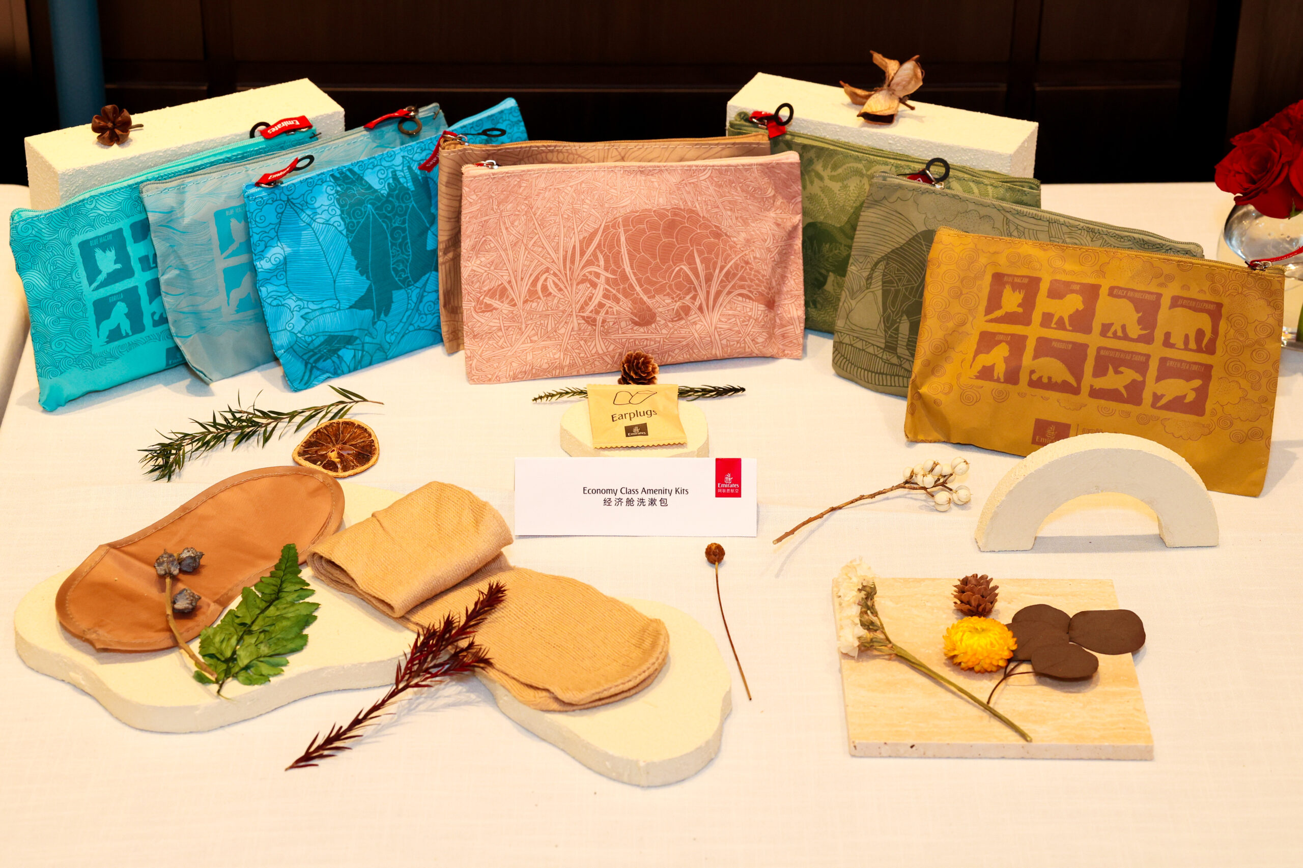 A display of various economy class amenity kits from an airline, featuring pouches in different colors and designs, along with earplugs, an eye mask, socks, and natural decorative elements such as leaves and dried flowers.