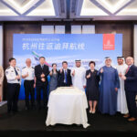 group of people, including airline staff and officials, stand in front of a backdrop featuring an Emirates airplane and Chinese text. They are posing for a photo with thumbs up gestures. Two Emirates flight attendants in uniform stand on either side of the group. A large cake is placed on a table in front of them.