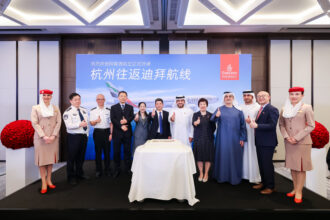 group of people, including airline staff and officials, stand in front of a backdrop featuring an Emirates airplane and Chinese text. They are posing for a photo with thumbs up gestures. Two Emirates flight attendants in uniform stand on either side of the group. A large cake is placed on a table in front of them.