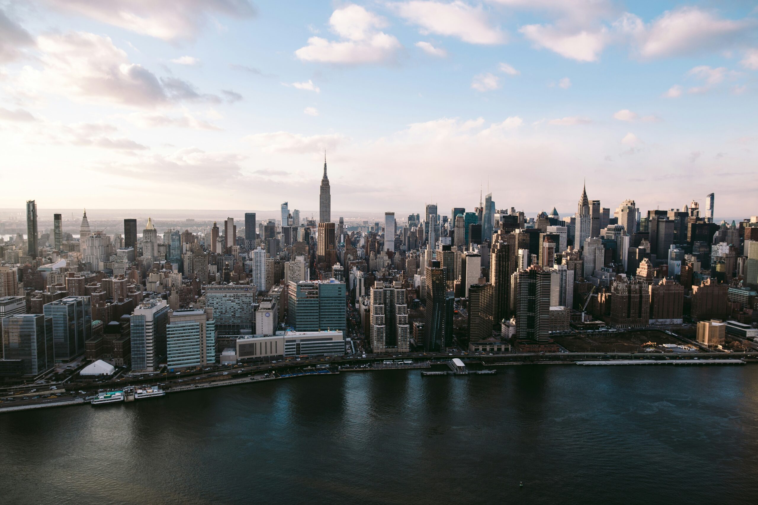 Aerial view of the New York City skyline during the day, featuring numerous skyscrapers including the iconic Empire State Building in the center and the Chrysler Building to its right. The East River is visible in the foreground.