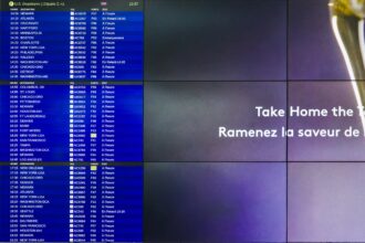 The image shows a digital departure board at an airport displaying flight information for U.S. departures. The board lists various destinations, flight numbers, gates, and statuses in both French and English. On the right side of the image, there is a partial advertisement with the text "Take Home the Taste" and "Ramenez la saveur de" in white letters on a purple background.
