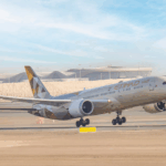 An Etihad Airways Boeing 787 Dreamliner aircraft is captured mid-takeoff from a runway at an airport. The plane's landing gear is still extended, and the background features a modern terminal building with a distinctive wavy roof design and a radar tower.