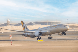 An Etihad Airways Boeing 787 Dreamliner aircraft is captured mid-takeoff from a runway at an airport. The plane's landing gear is still extended, and the background features a modern terminal building with a distinctive wavy roof design and a radar tower.