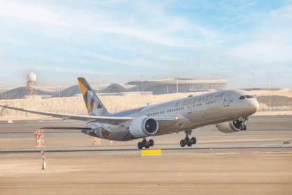 An Etihad Airways Boeing 787 Dreamliner aircraft is captured mid-takeoff from a runway at an airport. The plane's landing gear is still extended, and the background features a modern terminal building with a distinctive wavy roof design and a radar tower.