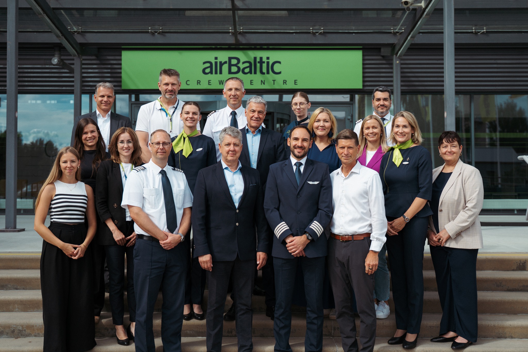 A group of airBaltic crew members and staff stand together in front of the airBaltic Crew Centre. The group includes pilots, flight attendants, and other staff members dressed in their respective uniforms.