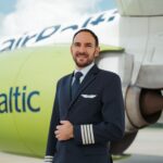 A pilot in a dark blue uniform with four stripes on the sleeve stands in front of an AirBaltic airplane. The airplane's engine is painted light green with the word "airBaltic" written on it in blue letters. The background shows part of the airplane's fuselage and wing, with a cloudy sky above.