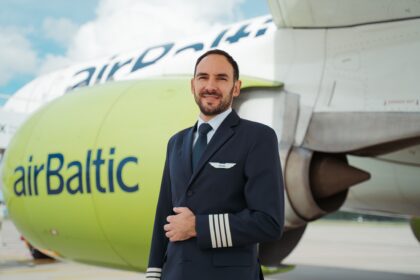 A pilot in a dark blue uniform with four stripes on the sleeve stands in front of an AirBaltic airplane. The airplane's engine is painted light green with the word "airBaltic" written on it in blue letters. The background shows part of the airplane's fuselage and wing, with a cloudy sky above.