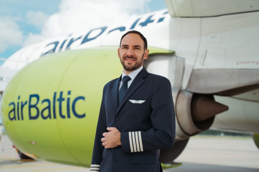 A pilot in a dark blue uniform with four stripes on the sleeve stands in front of an AirBaltic airplane. The airplane's engine is painted light green with the word "airBaltic" written on it in blue letters. The background shows part of the airplane's fuselage and wing, with a cloudy sky above.