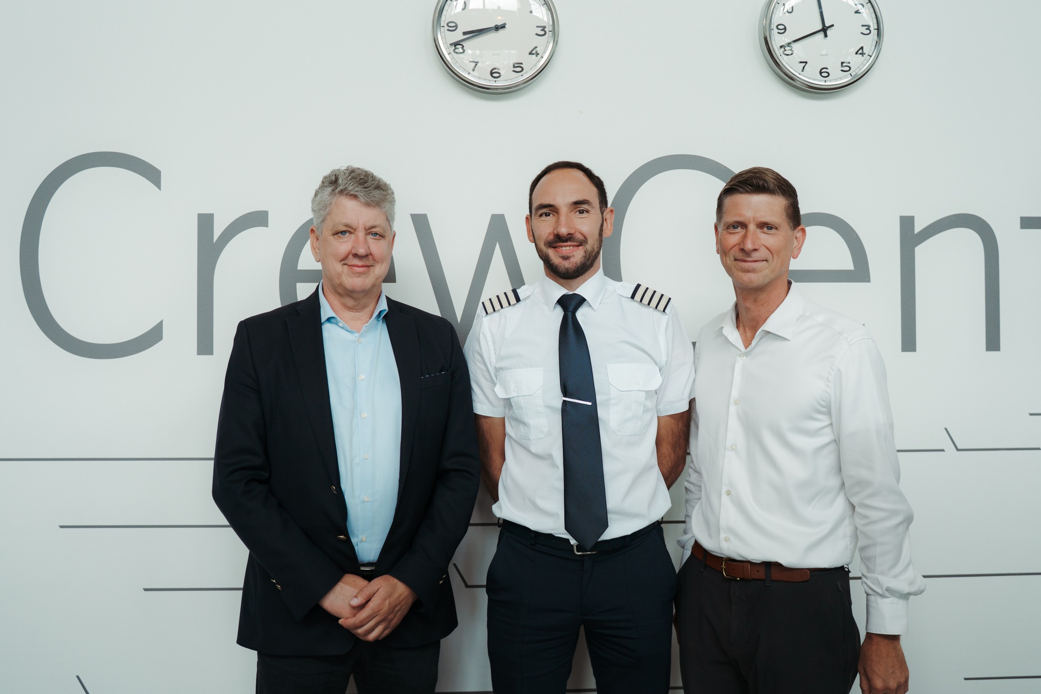 Three men standing in front of a wall with the word "Crew Center" partially visible. The man on the left is wearing a dark suit, the man in the middle is dressed in a pilot's uniform with epaulets and a tie, and the man on the right is wearing a white dress shirt. Two clocks are mounted on the wall above them, showing different times.