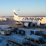 A Finnair airplane is parked at a snowy airport gate, with ground service vehicles and equipment surrounding it. Another Finnair aircraft can be seen in the background on the tarmac. The sky is clear and blue, indicating a cold but sunny day.