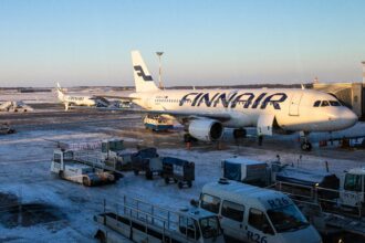 A Finnair airplane is parked at a snowy airport gate, with ground service vehicles and equipment surrounding it. Another Finnair aircraft can be seen in the background on the tarmac. The sky is clear and blue, indicating a cold but sunny day.