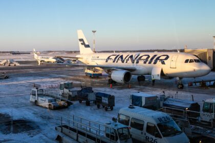 A Finnair airplane is parked at a snowy airport gate, with ground service vehicles and equipment surrounding it. Another Finnair aircraft can be seen in the background on the tarmac. The sky is clear and blue, indicating a cold but sunny day.