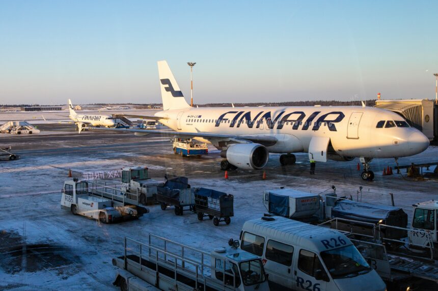 A Finnair airplane is parked at a snowy airport gate, with ground service vehicles and equipment surrounding it. Another Finnair aircraft can be seen in the background on the tarmac. The sky is clear and blue, indicating a cold but sunny day.