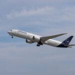 A Lufthansa Boeing 787 Dreamliner aircraft is captured mid-flight against a backdrop of a partly cloudy sky. The airplane, painted in the airline's white and blue livery with the Lufthansa logo on the tail, is ascending with its landing gear partially retracted.