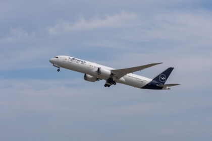 A Lufthansa Boeing 787 Dreamliner aircraft is captured mid-flight against a backdrop of a partly cloudy sky. The airplane, painted in the airline's white and blue livery with the Lufthansa logo on the tail, is ascending with its landing gear partially retracted.