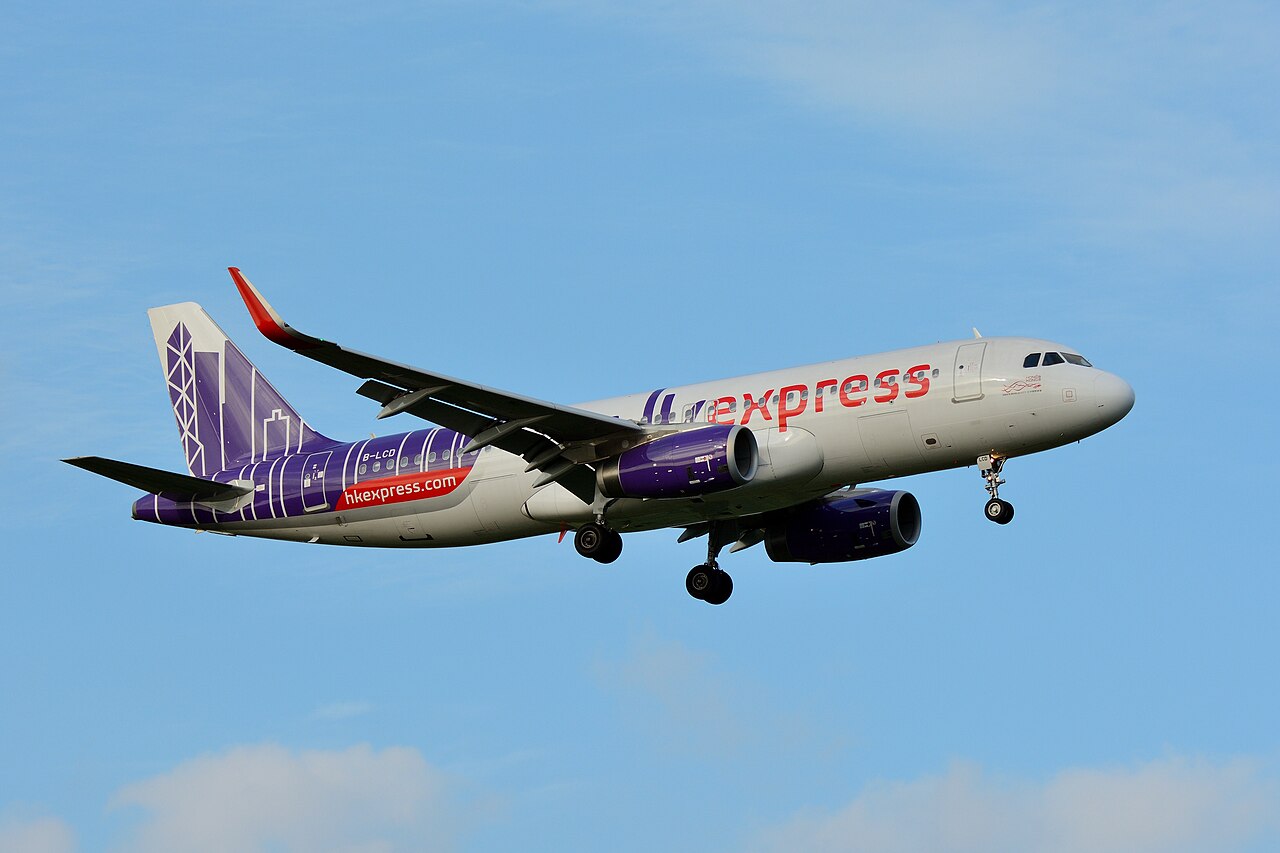 A HK Express aircraft with registration number B-LCD is captured in mid-air during its landing approach. The aeroplane features a white fuselage with "HK Express" branding in red and purple, and a distinctive purple tail design with geometric patterns. The landing gear is deployed, and the sky is clear blue.
