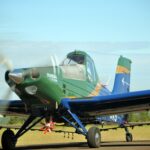 A green and blue agricultural aircraft with the name "IPANEMA 203" on its side is parked on a runway. The propeller is spinning, creating a blurred effect. The aircraft has a single cockpit and is equipped with spray nozzles underneath its wings.