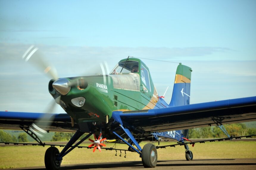 A green and blue agricultural aircraft with the name "IPANEMA 203" on its side is parked on a runway. The propeller is spinning, creating a blurred effect. The aircraft has a single cockpit and is equipped with spray nozzles underneath its wings.