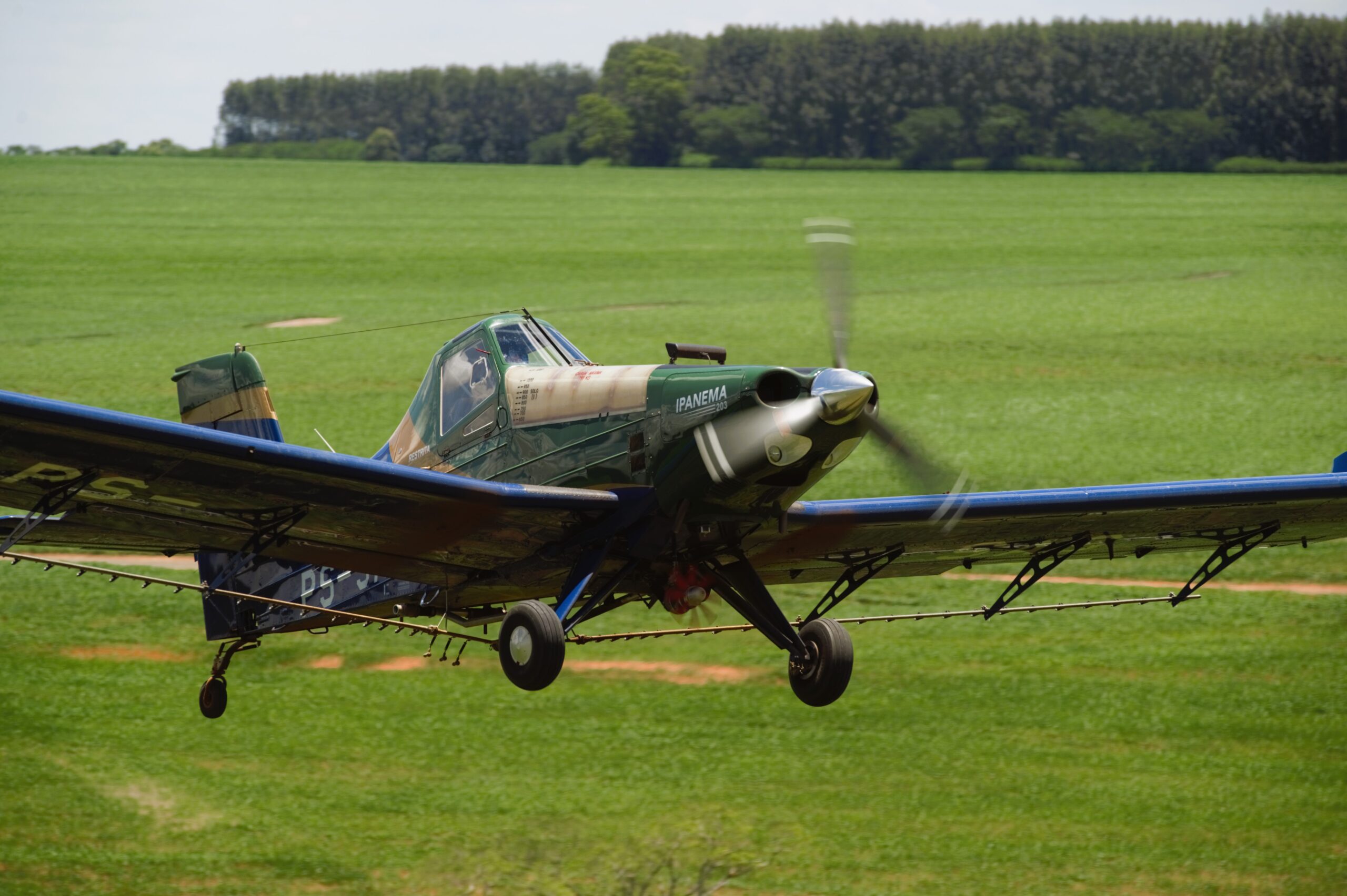 A crop duster airplane, labeled "IPANEMA" on the nose, flying low over a vast green agricultural field with a blurred propeller indicating motion. The background features a line of trees under a partly cloudy sky.