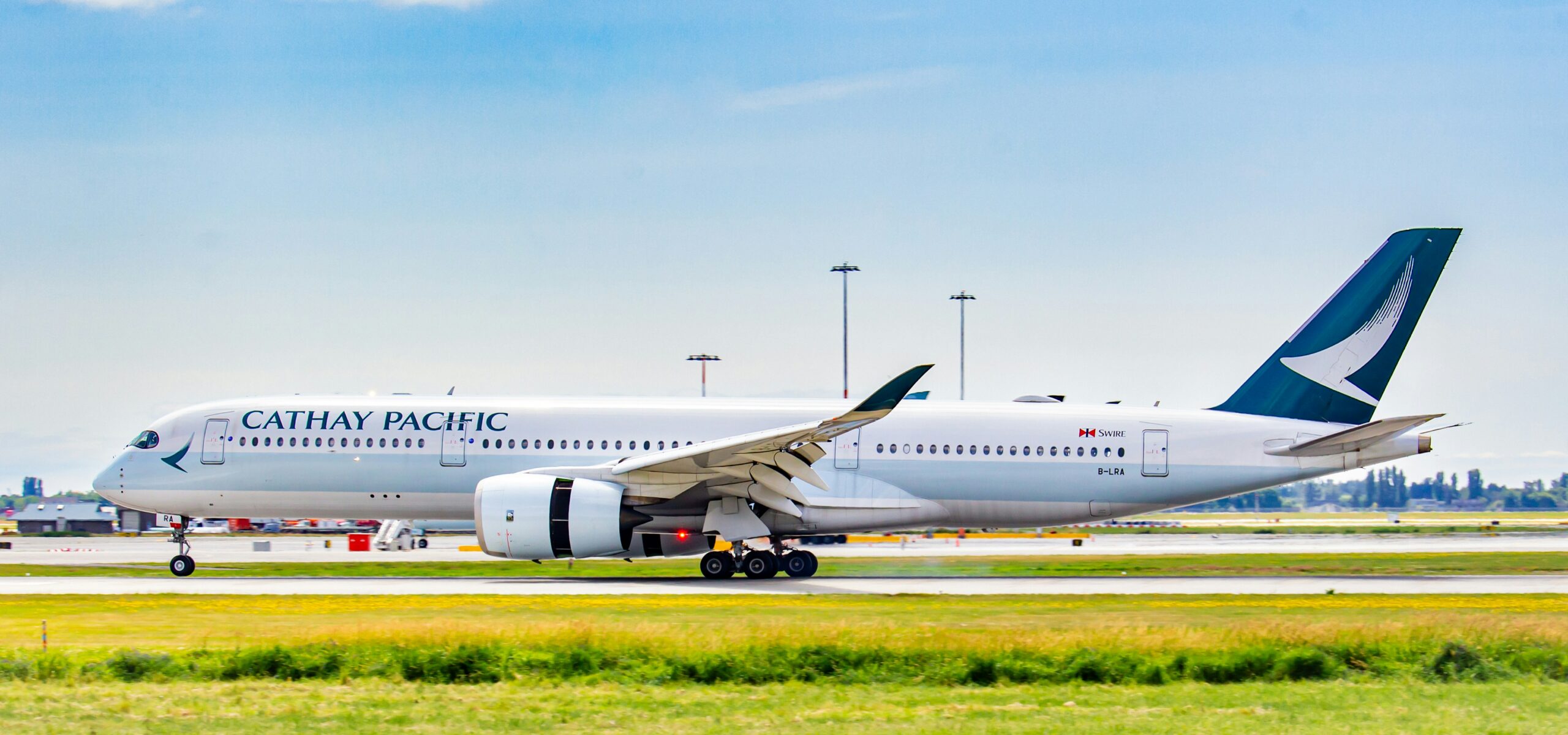 A Cathay Pacific aeroplane is captured taxiing on a runway at an airport. The aircraft is white with the airline's name "CATHAY PACIFIC" written in large letters on the fuselage. The tail fin features the airline's logo, a stylised brush wing, in green and white. The registration number "B-LRA" is visible near the rear of the plane.