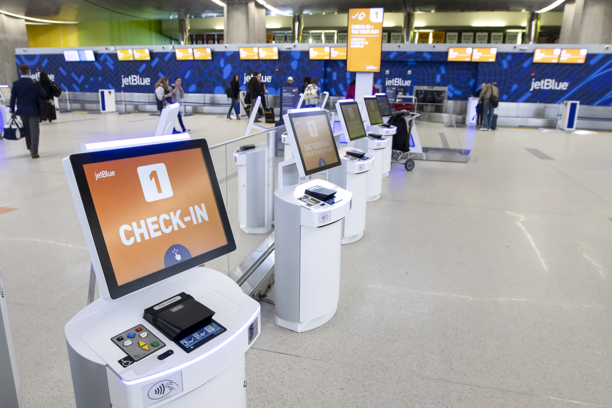 A row of self-service check-in kiosks at a JetBlue terminal in an airport. The kiosks have touch screens displaying "CHECK-IN" with the number 1 and the JetBlue logo. In the background, there are passengers walking and checking in at counters with blue JetBlue branding.