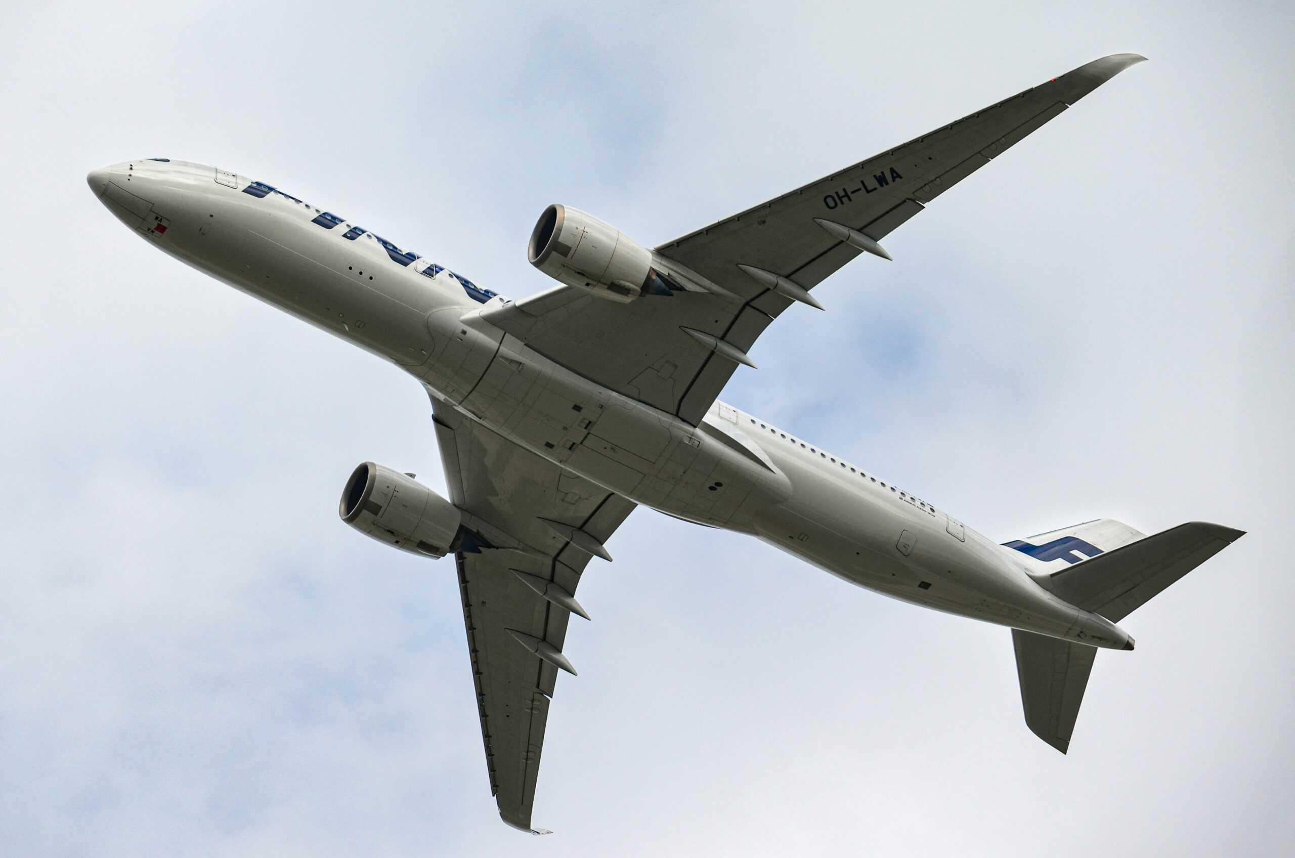 A Finnair airplane, with registration OH-LWA, is captured from below as it flies through a partly cloudy sky. The aircraft's underbelly and wings are clearly visible, showcasing the twin engines mounted under the wings.